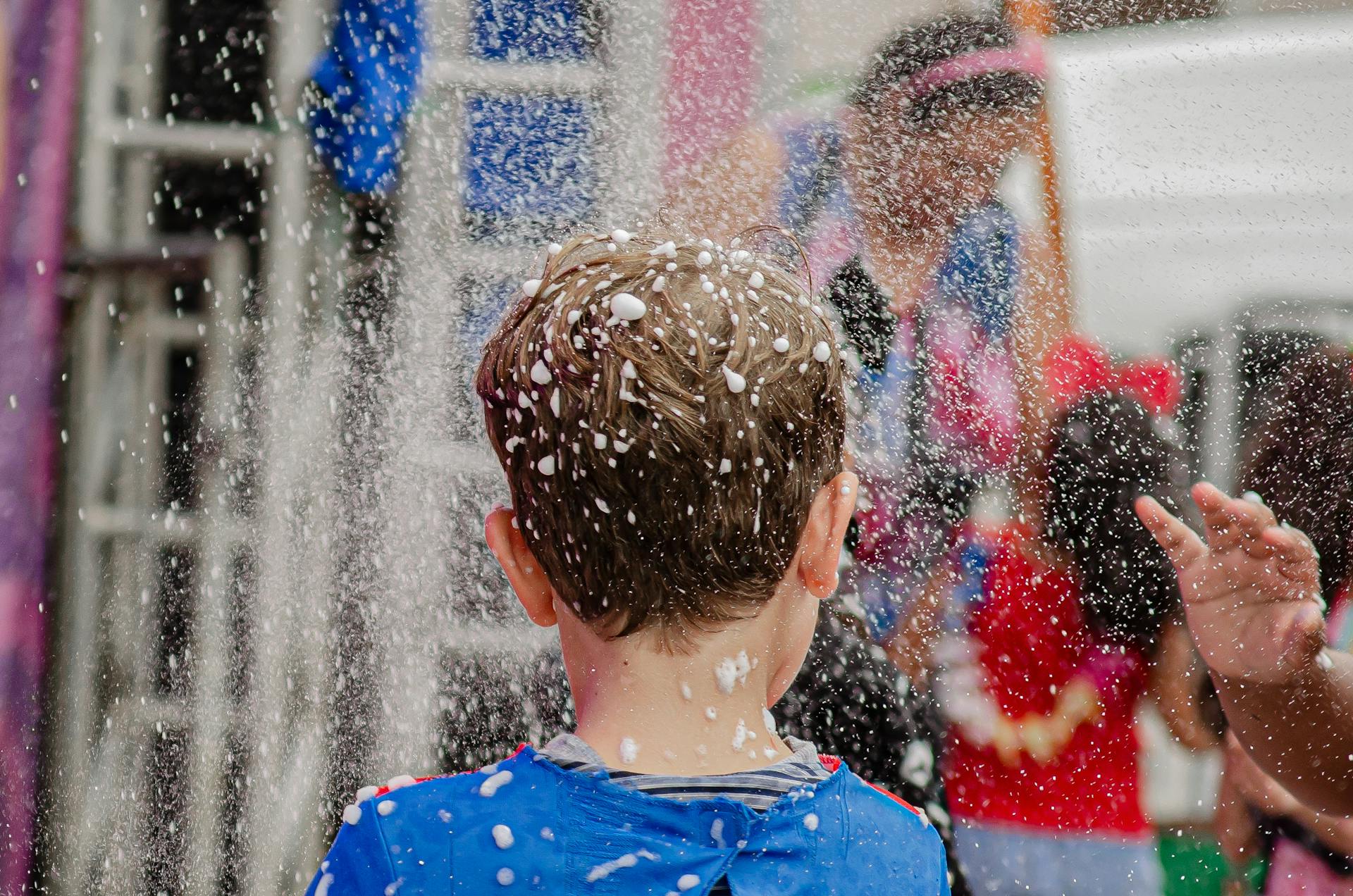 A young child with brown hair plays joyfully in foam spray during an outdoor party.