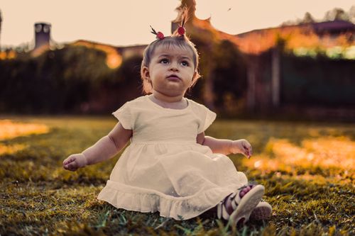 Free Cute toddler sitting on grass in a white dress under warm sunlight, capturing innocence and joy. Stock Photo