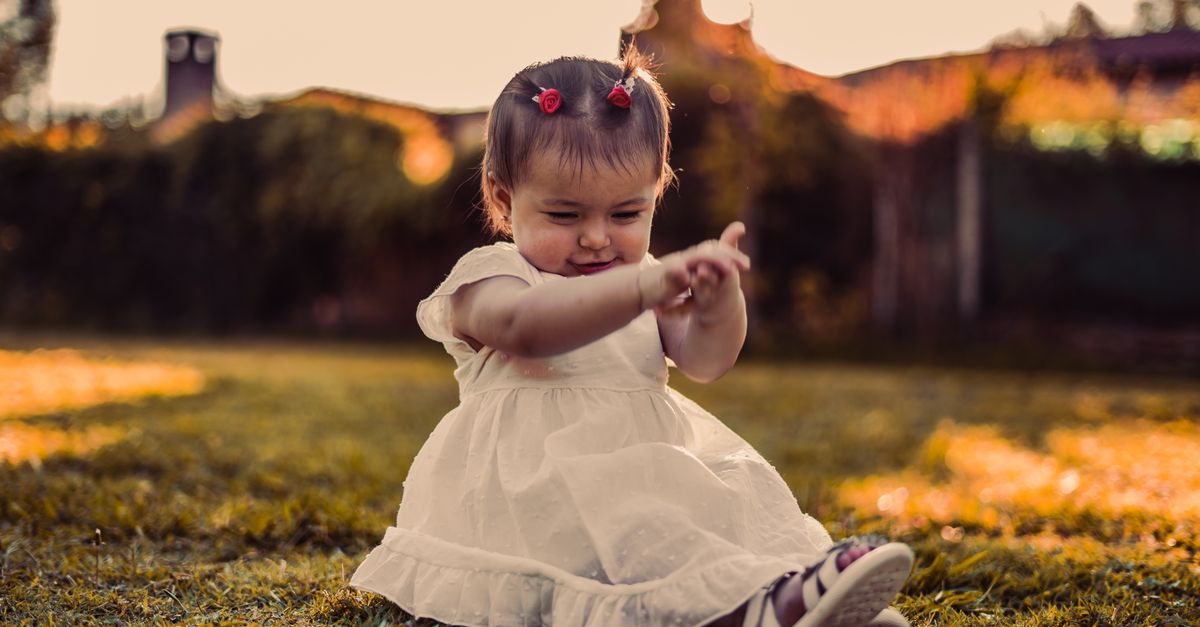Adorable baby girl in white dress playing on grass during sunset, capturing joy and innocence.