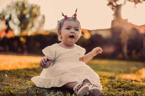 A Toddler in a White Dress Sitting on the Grass