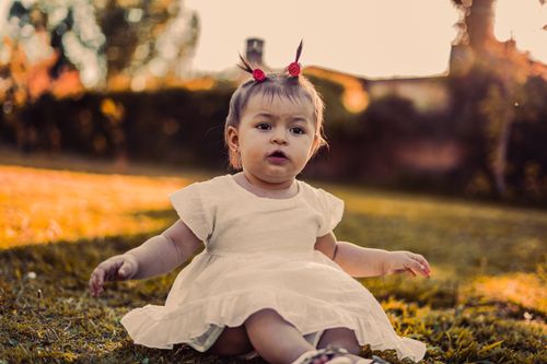 Free Charming baby girl with pigtails sitting on grass at sunset in a park. Stock Photo