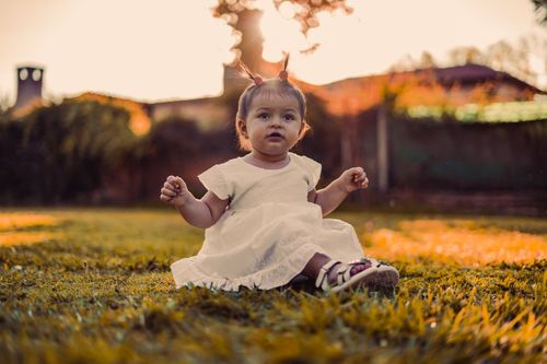 Free Charming baby girl in white dress sitting on grassy field at sunset, capturing a warm, serene moment outdoors. Stock Photo