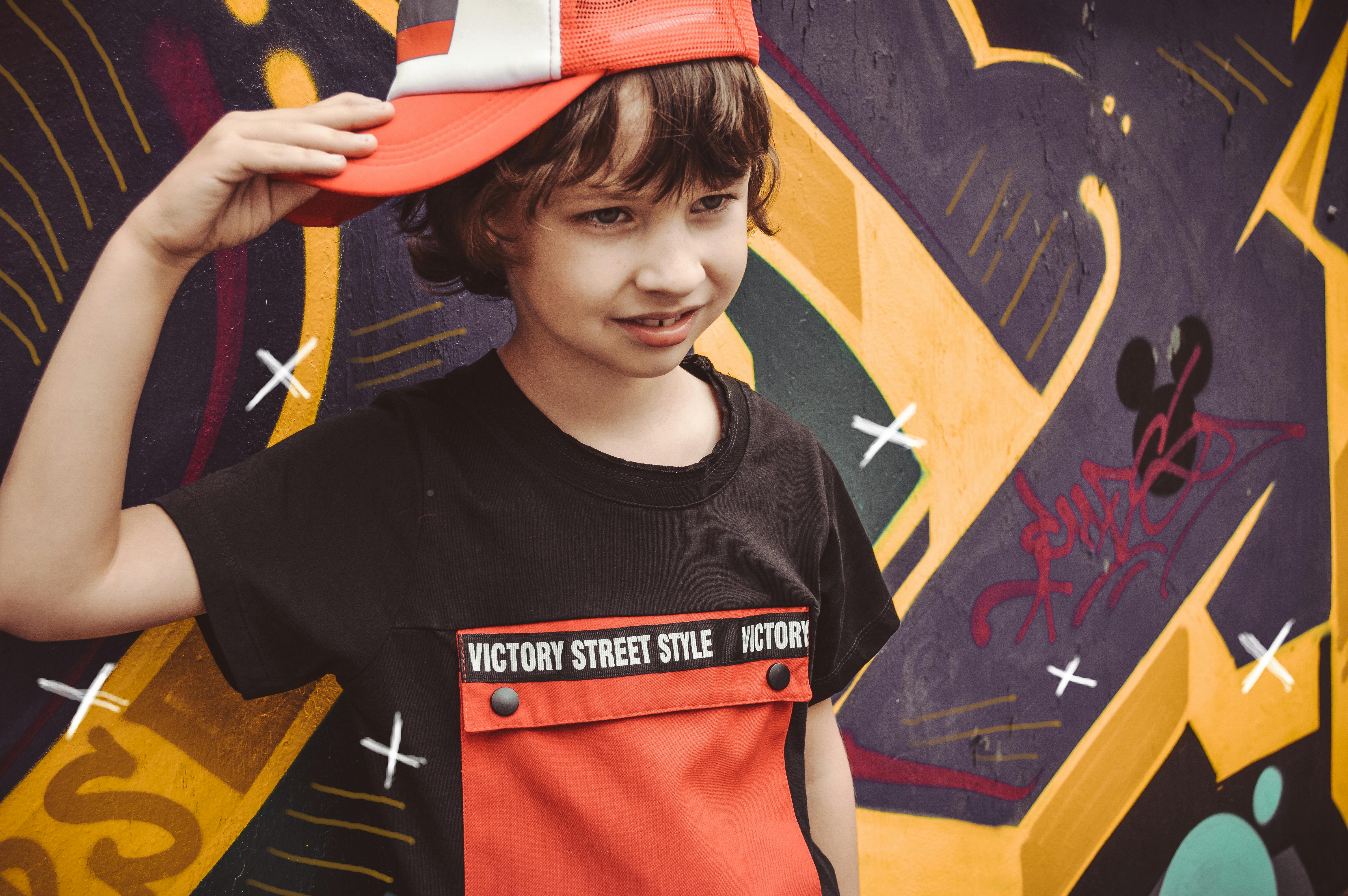 A cheerful child wearing a red cap poses near a vibrant graffiti wall.