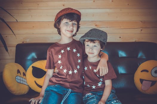 两个男孩穿着相配的明星衬衫和帽子，坐在沙发上微笑，周围堆满了表情符号枕头