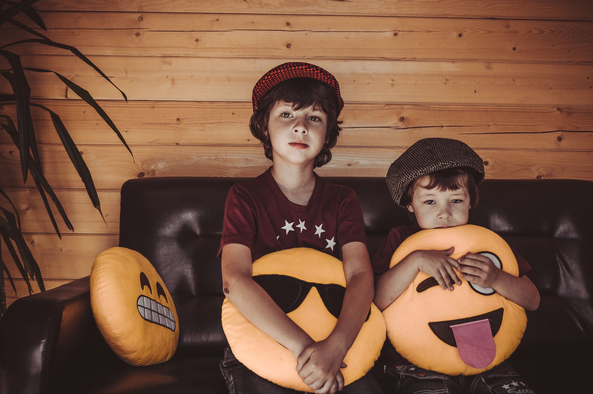 Two boys in matching hats sit indoors holding emoji pillows, expressing playfulness.