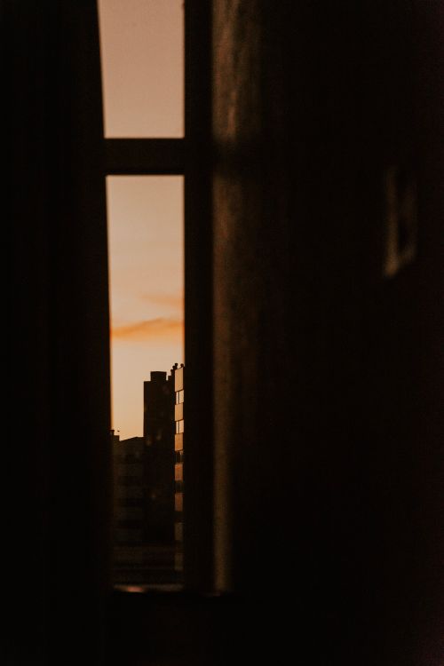Free A dramatic view of urban buildings silhouetted against a sunset through a window. Stock Photo