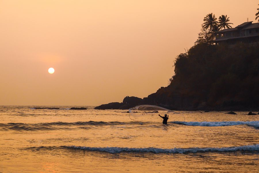 Silhouette of a fisherman casting a net against a stunning sunset backdrop on the coastline.
