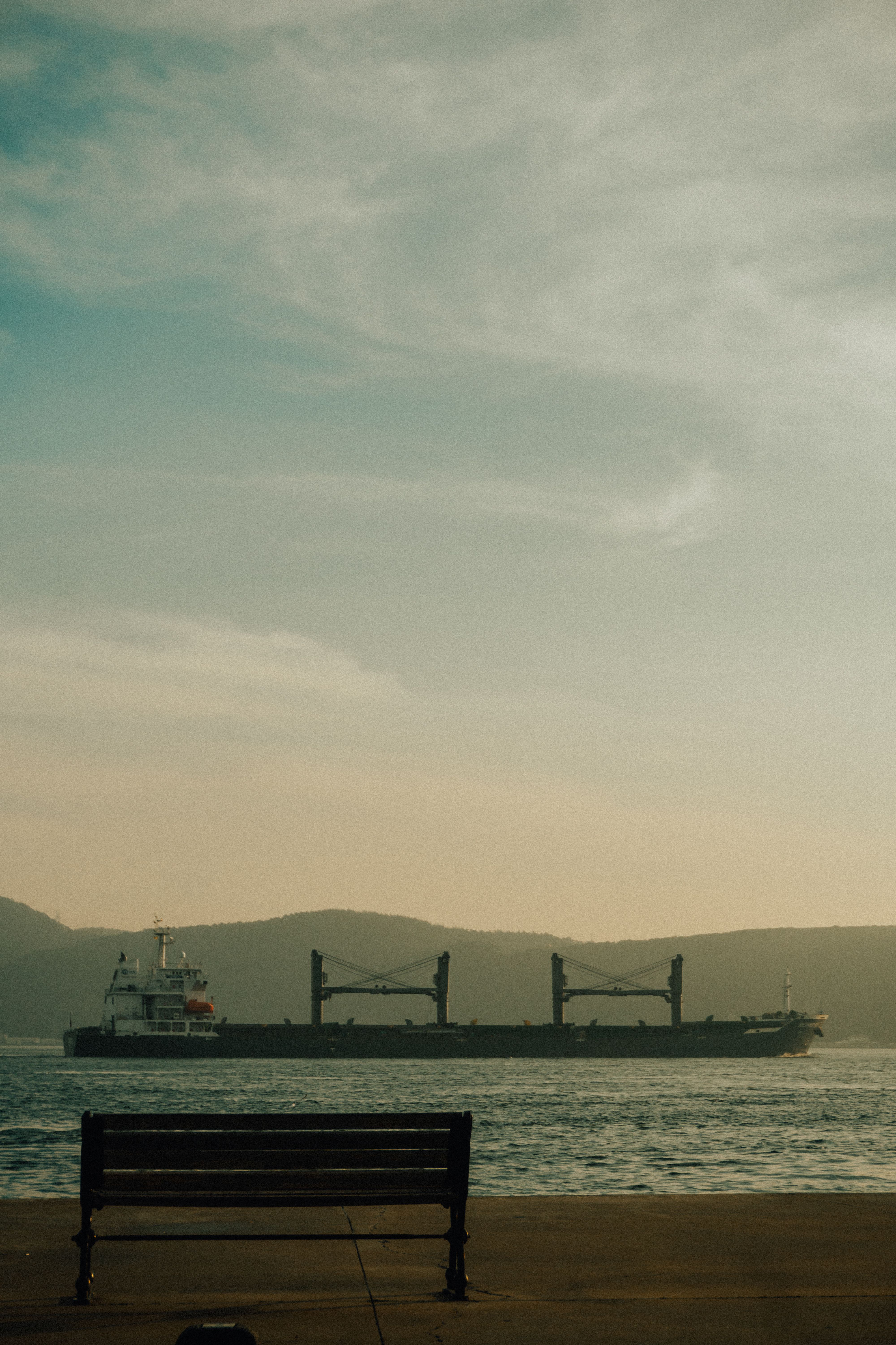 Peaceful scene of a ship sailing past an empty bench by the waterfront under a blue sky.