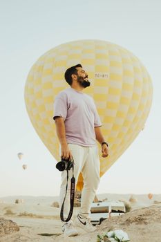 A man with a camera stands before a hot air balloon in the scenic landscape of Cappadocia, Türkiye.