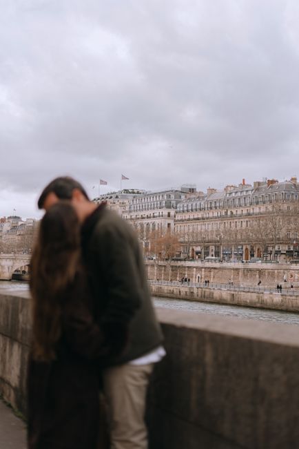 A couple shares a tender moment along the Seine River, with classic Parisian architecture in the backdrop.