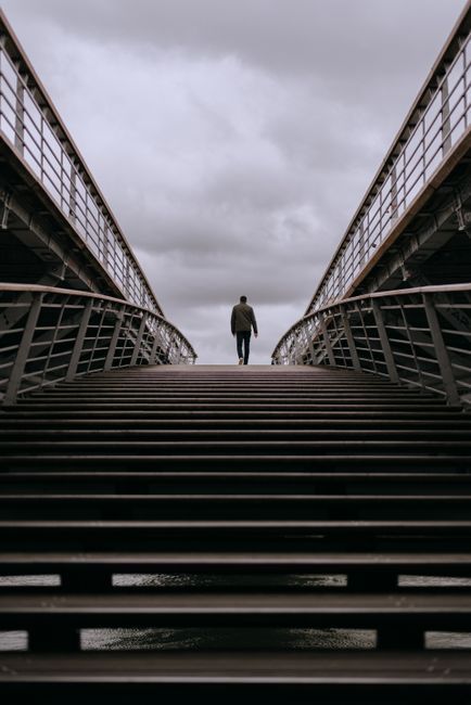 A solitary man walks across a contemporary steel bridge in Paris under cloudy skies.