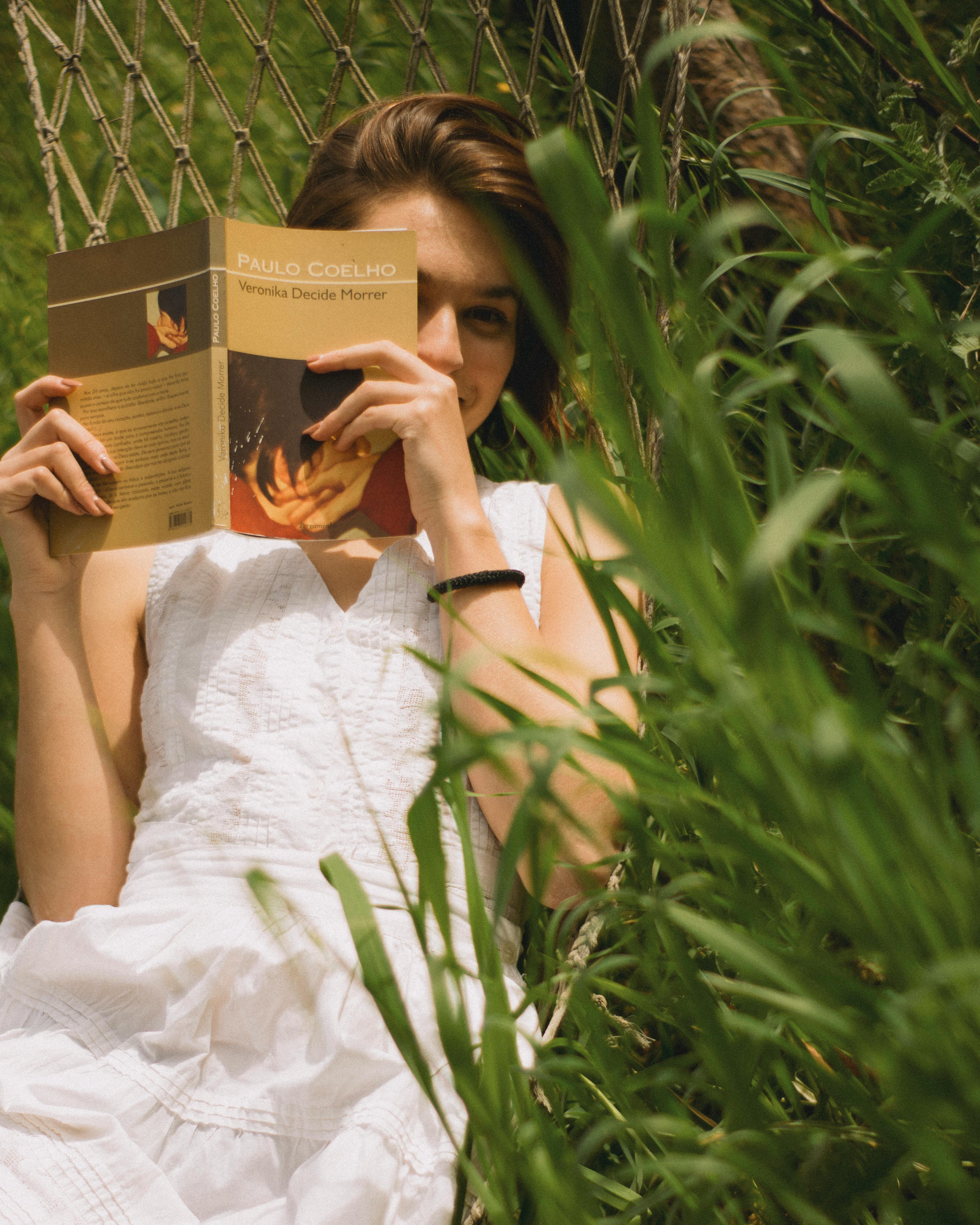 Young woman in a white dress reading a book on a hammock in a sunny grass field.