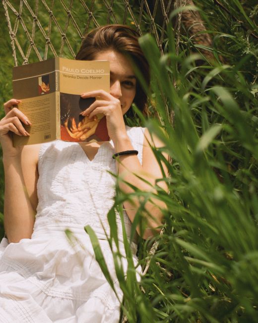 Young woman in a white dress reading a book on a hammock in a sunny grass field.