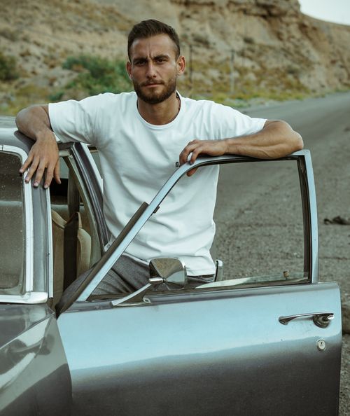 Free A man in a white t-shirt leaning against an open car door on a desert highway. Stock Photo