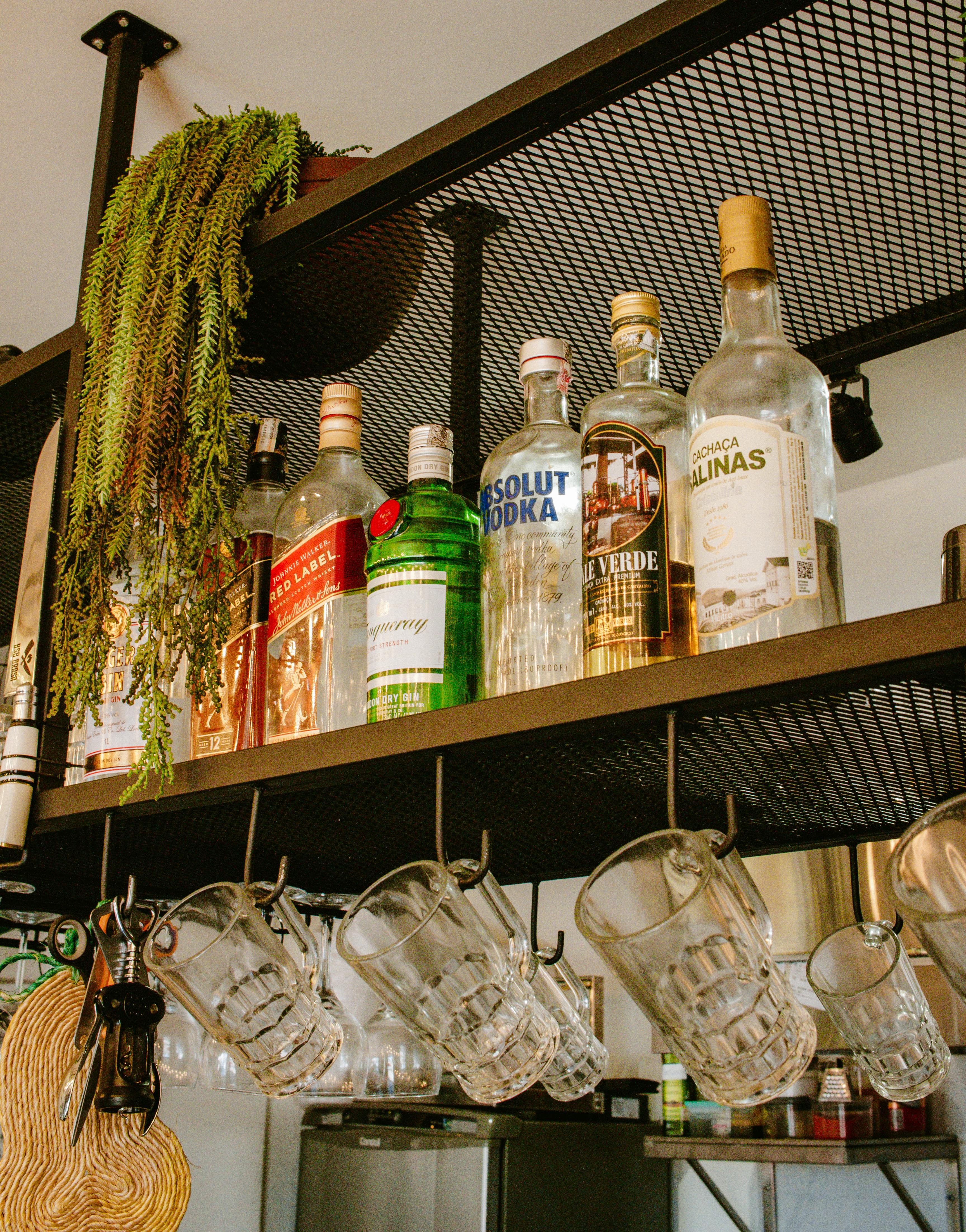 A collection of various liquor bottles and glassware on a bar shelf with hanging mugs.