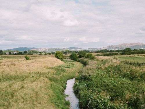 Free Scenic rural landscape in Rhuddlan, Wales, showcasing a gentle brook and lush fields under a cloudy sky. Stock Photo