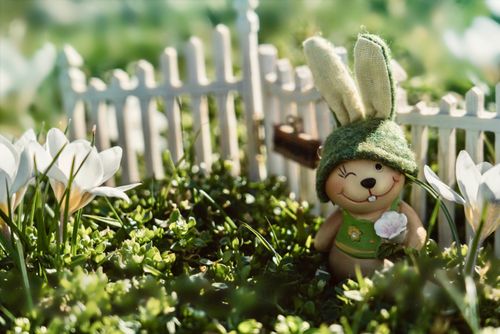 Free Adorable bunny figurine in a sunny garden surrounded by blooming flowers and a white picket fence. Stock Photo