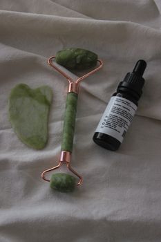 Flat lay of jade roller, gua sha, and serum bottle on soft fabric background.