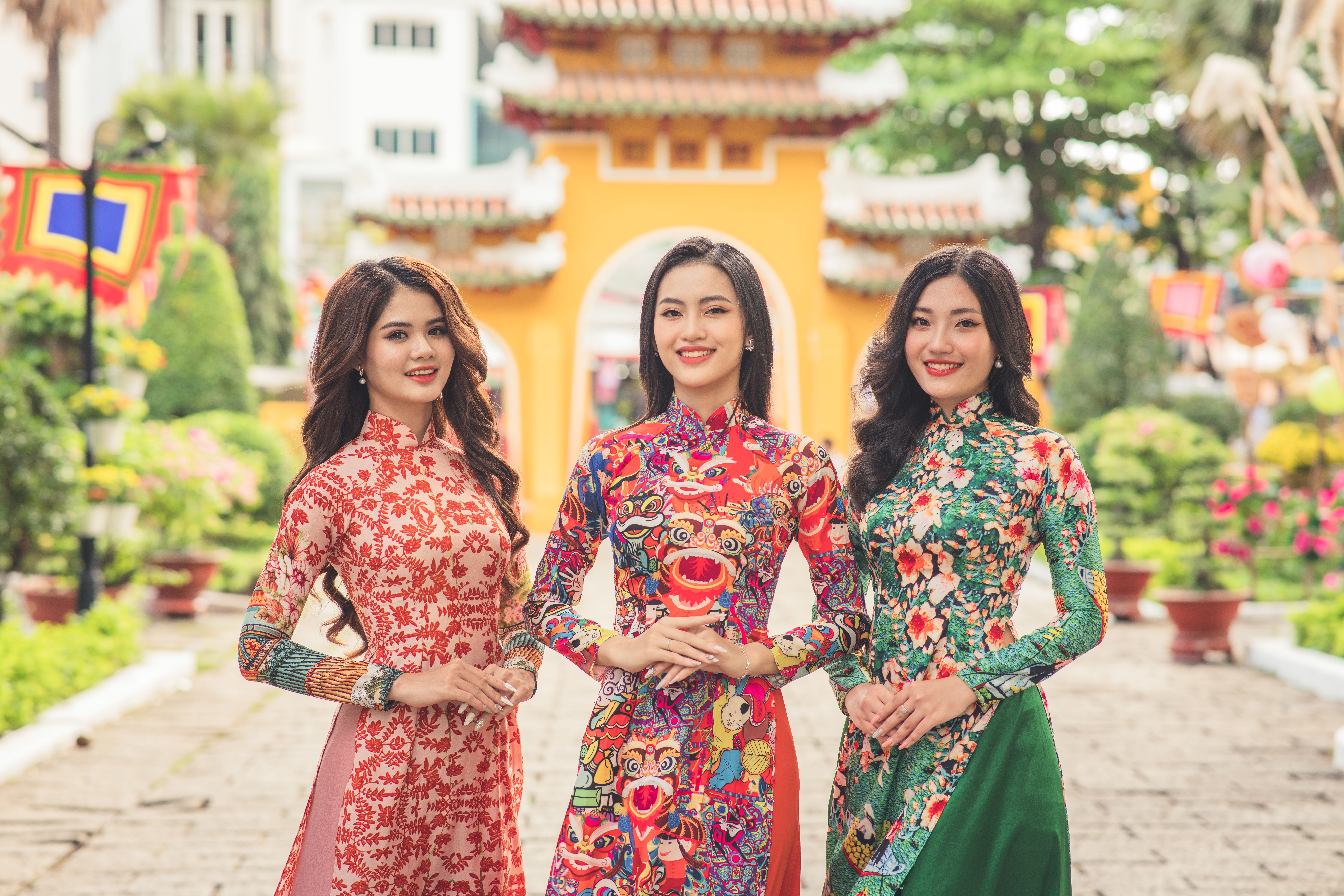 Three Asian women in colorful traditional dresses smiling in an urban setting with a cultural backdrop.