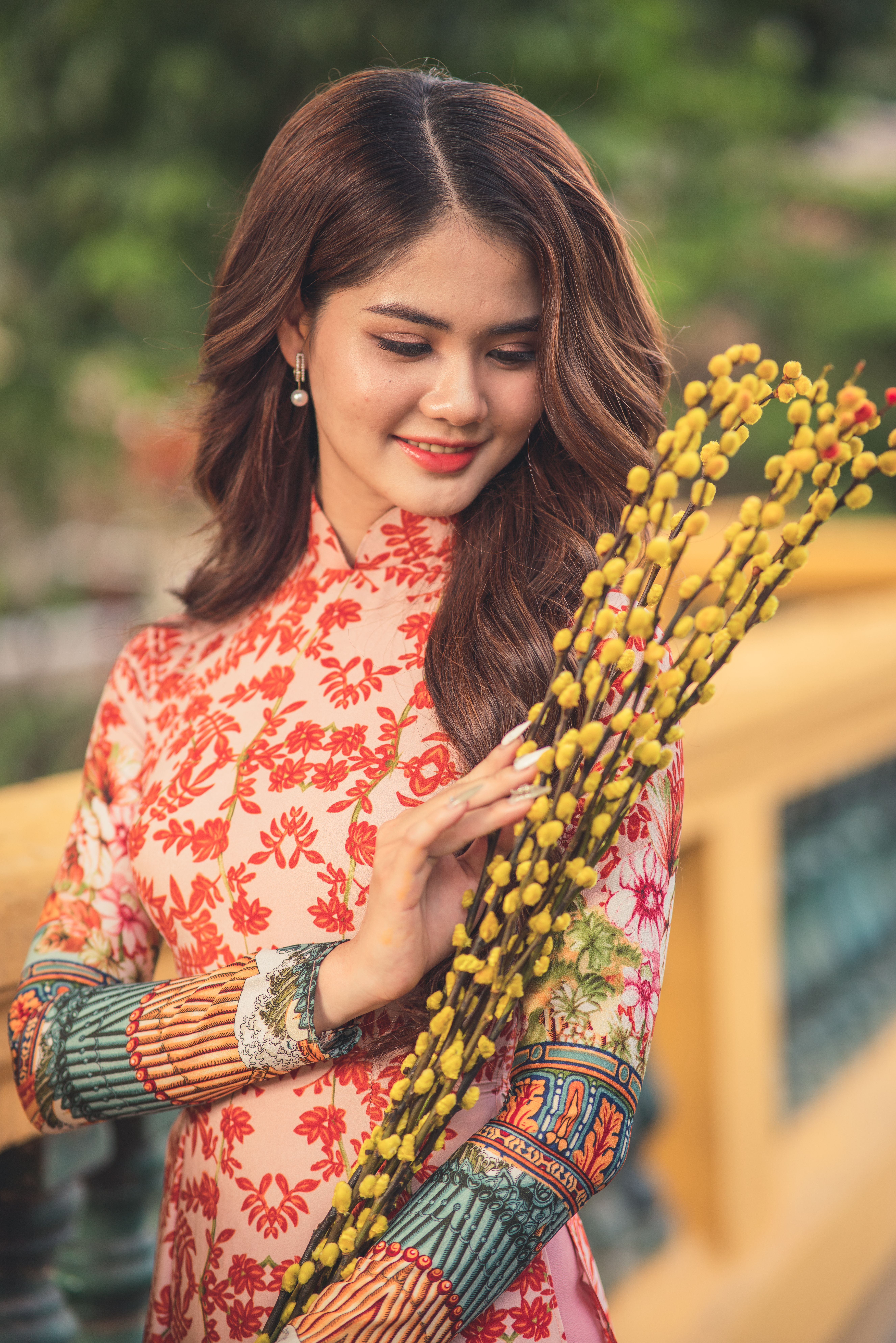 Beautiful woman in floral dress holding yellow catkins on a picturesque bridge.