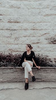 Stylish woman posing seated against a textured brick wall outdoors.