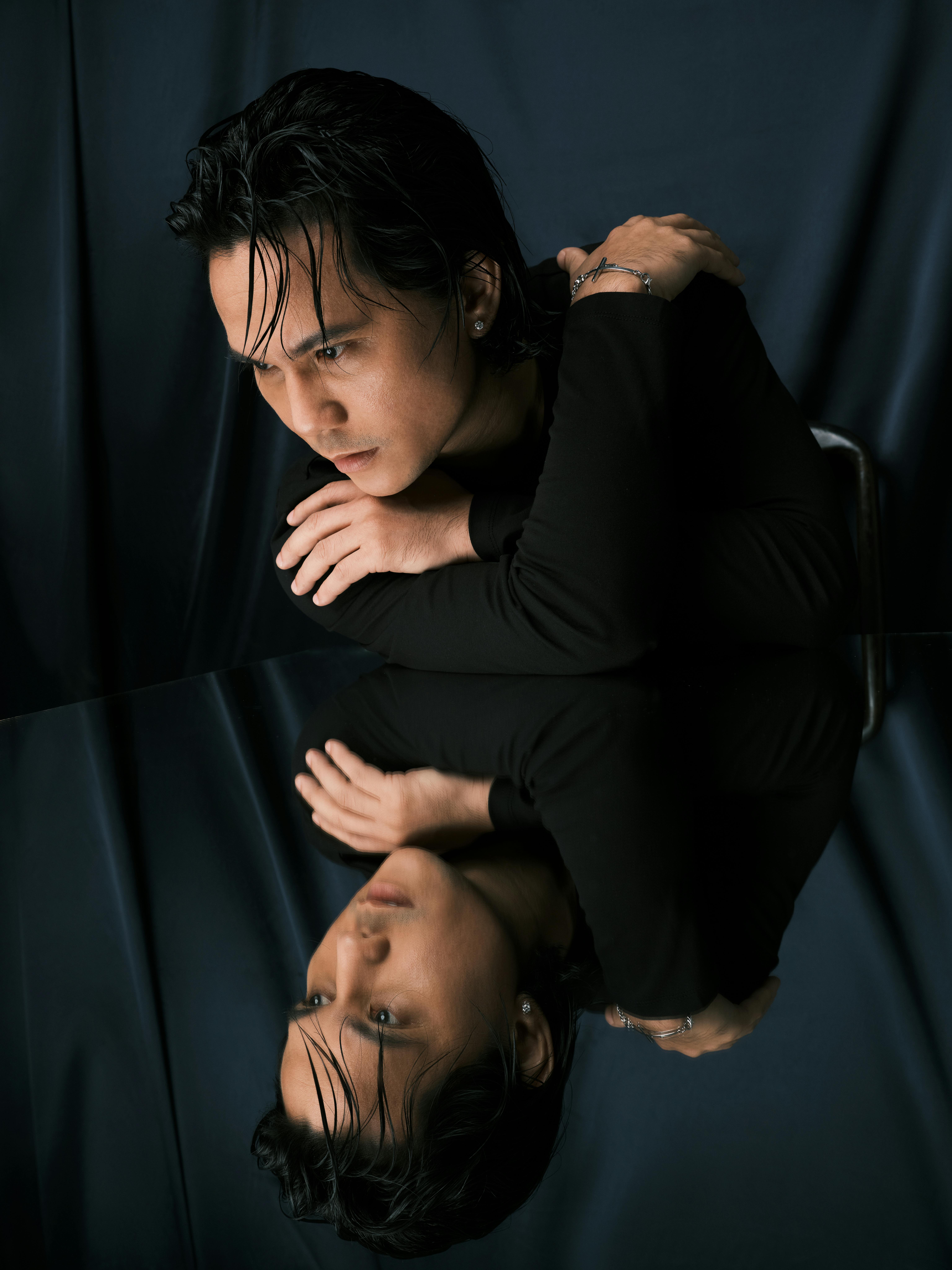 Contemplative Asian man in black with artistic mirror reflection. Studio lighting.