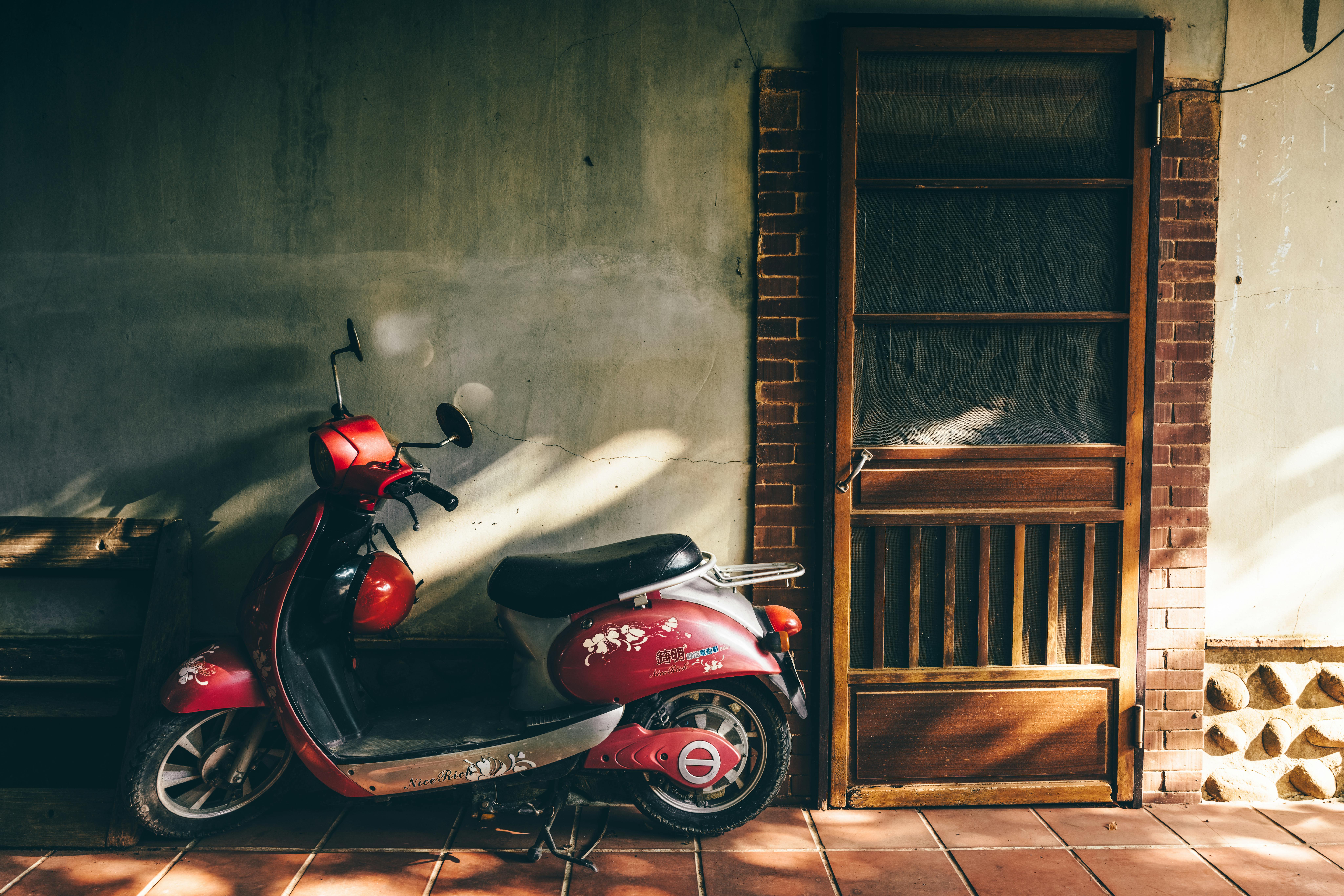 A vintage red scooter parked next to a wooden door in warm sunlight, casting shadows on a rustic wall.