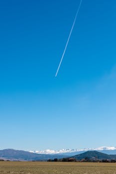 A clear blue sky with a jet contrail above distant snowy mountains and a grassy field.