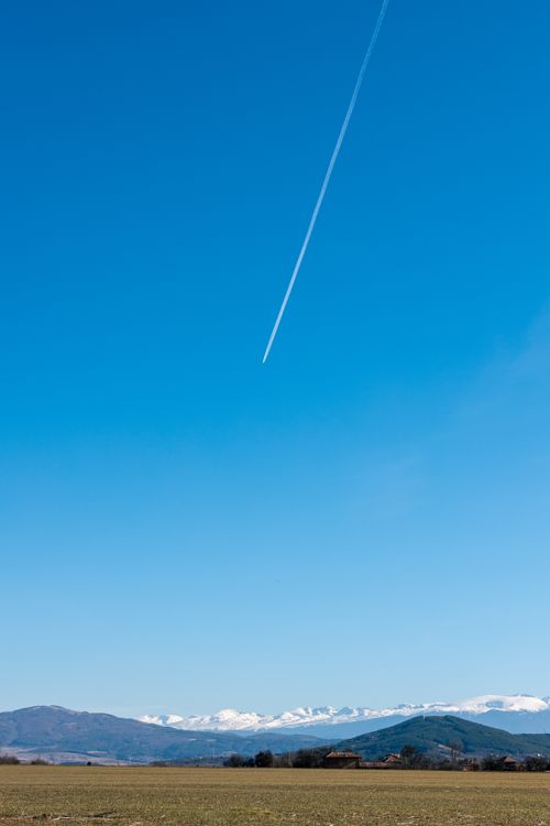 Free A clear blue sky with a jet contrail above distant snowy mountains and a grassy field. Stock Photo