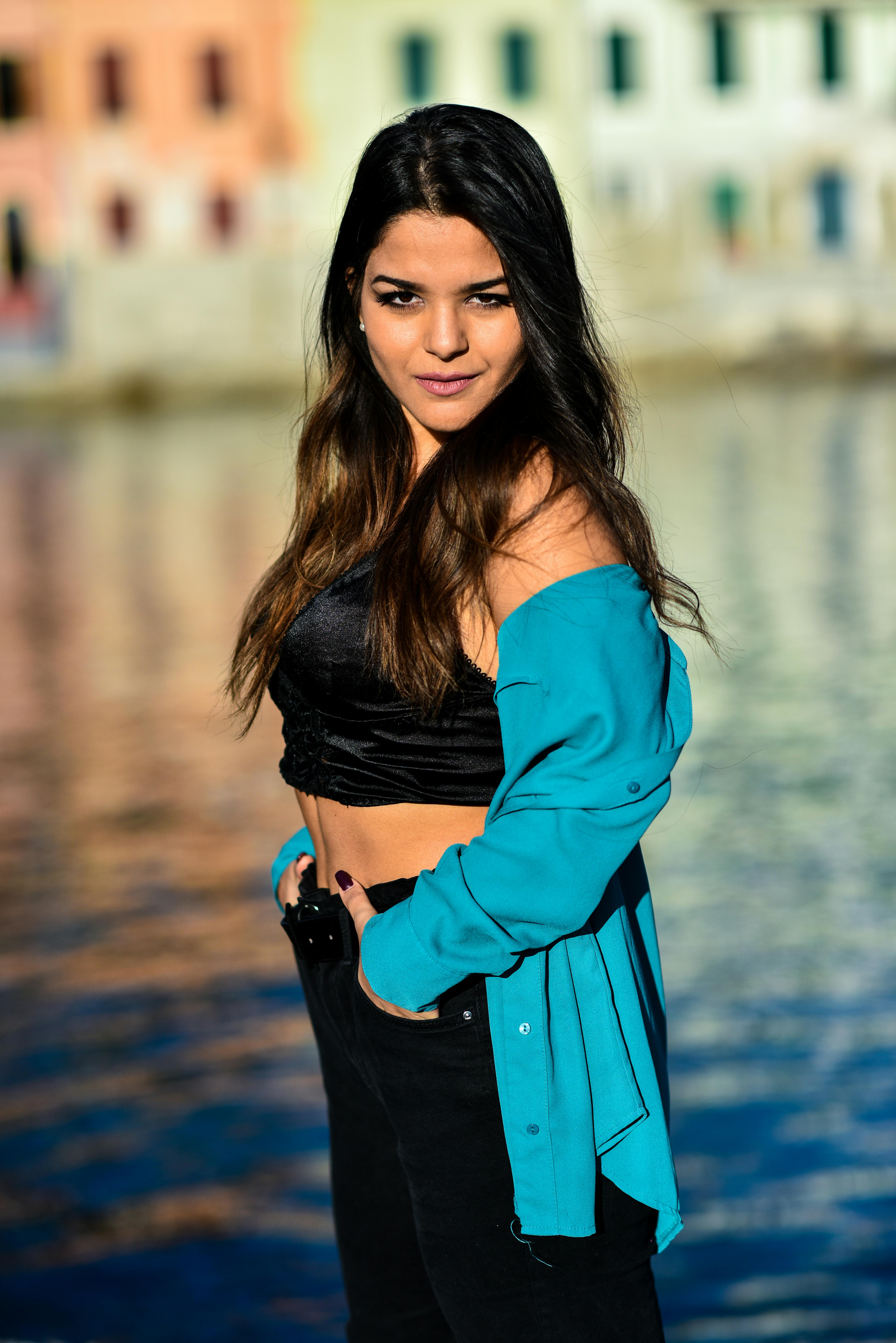 A fashionable woman poses confidently on a sunny day by a vibrant waterfront cityscape.
