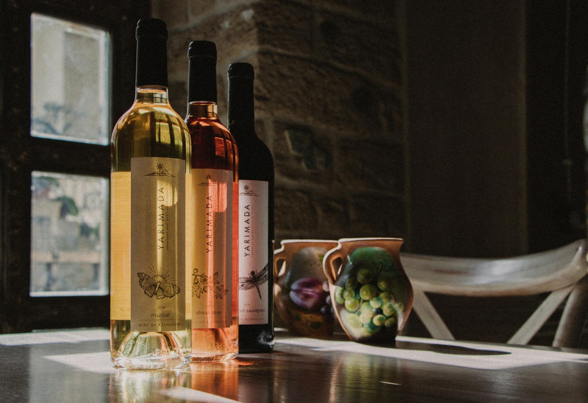 Three wine bottles with artistic labels on a sunlit wooden table beside painted jugs.