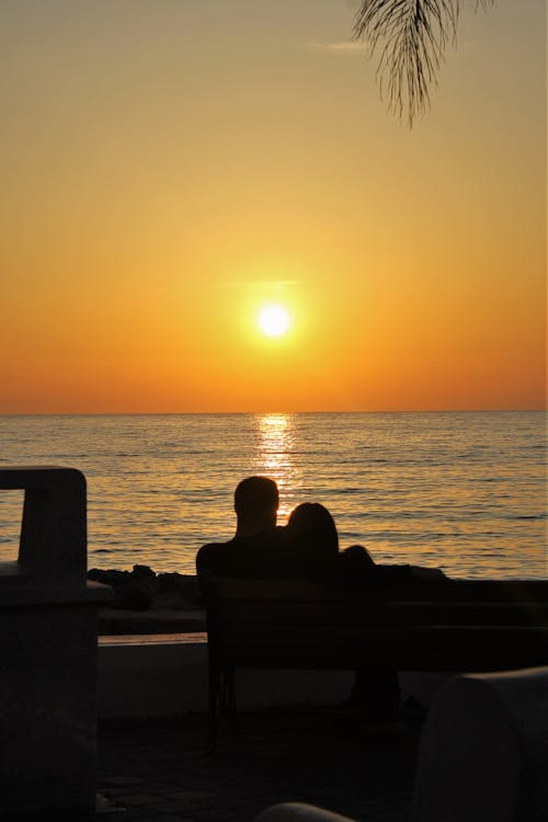 Silhouetted Couple Sitting on a Bench on the Shore, Watching the Sunset 