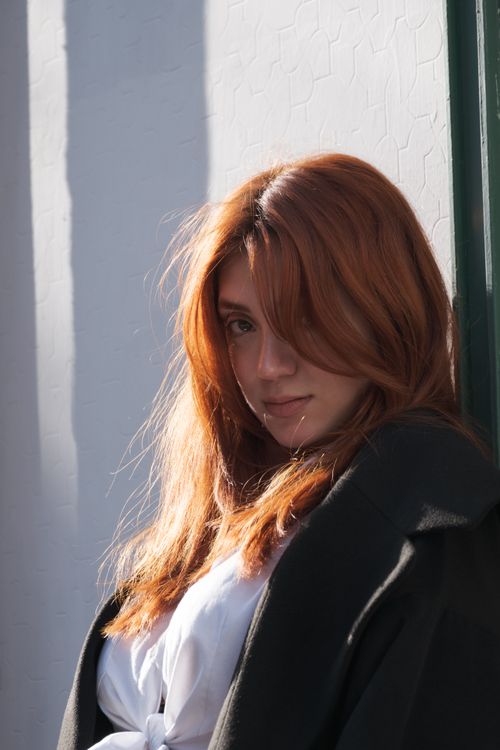 Free A woman with red hair stands against a wall, illuminated by sunlight. Stock Photo