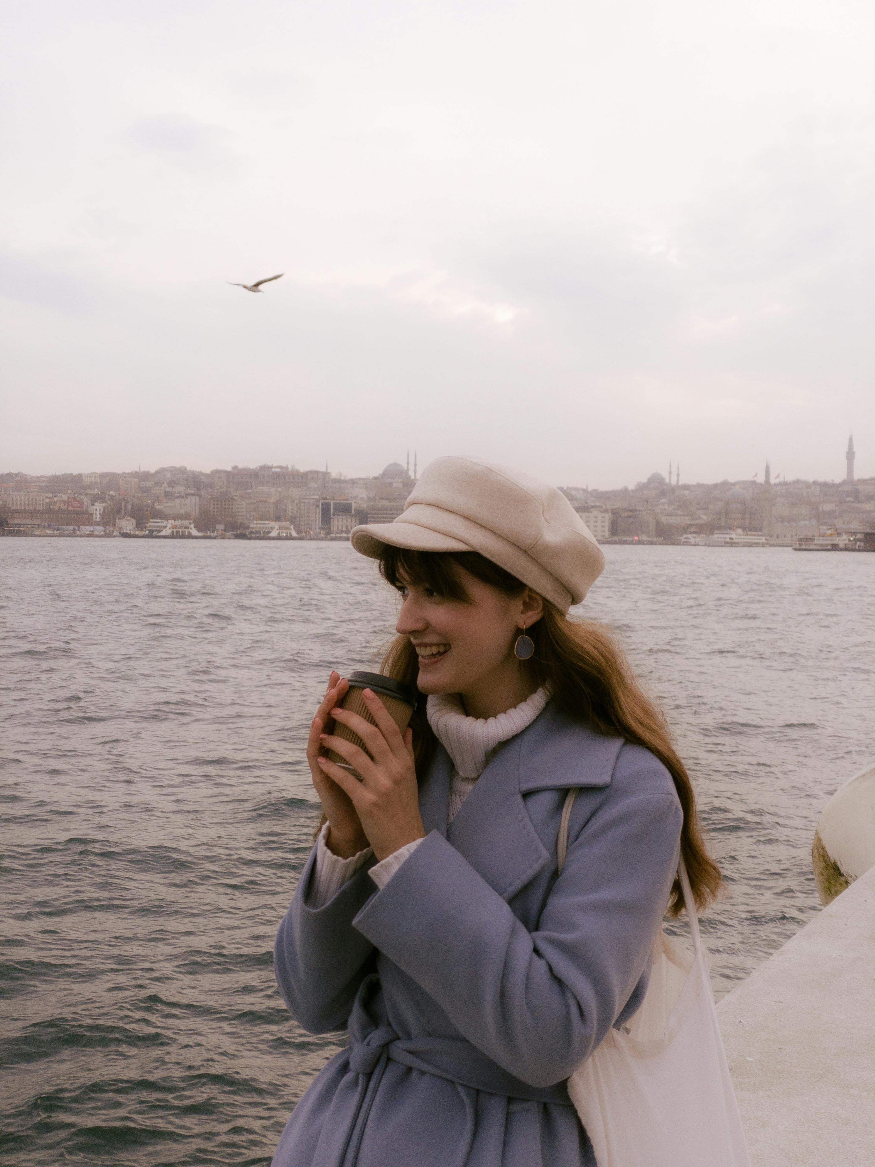 穿着外套和帽子的女人微笑着，在海边拿着咖啡