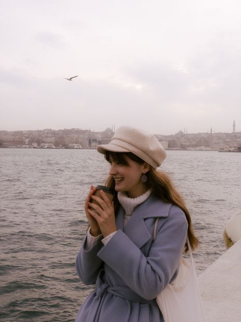 Woman in a coat and hat smiling while holding a coffee by the sea.
