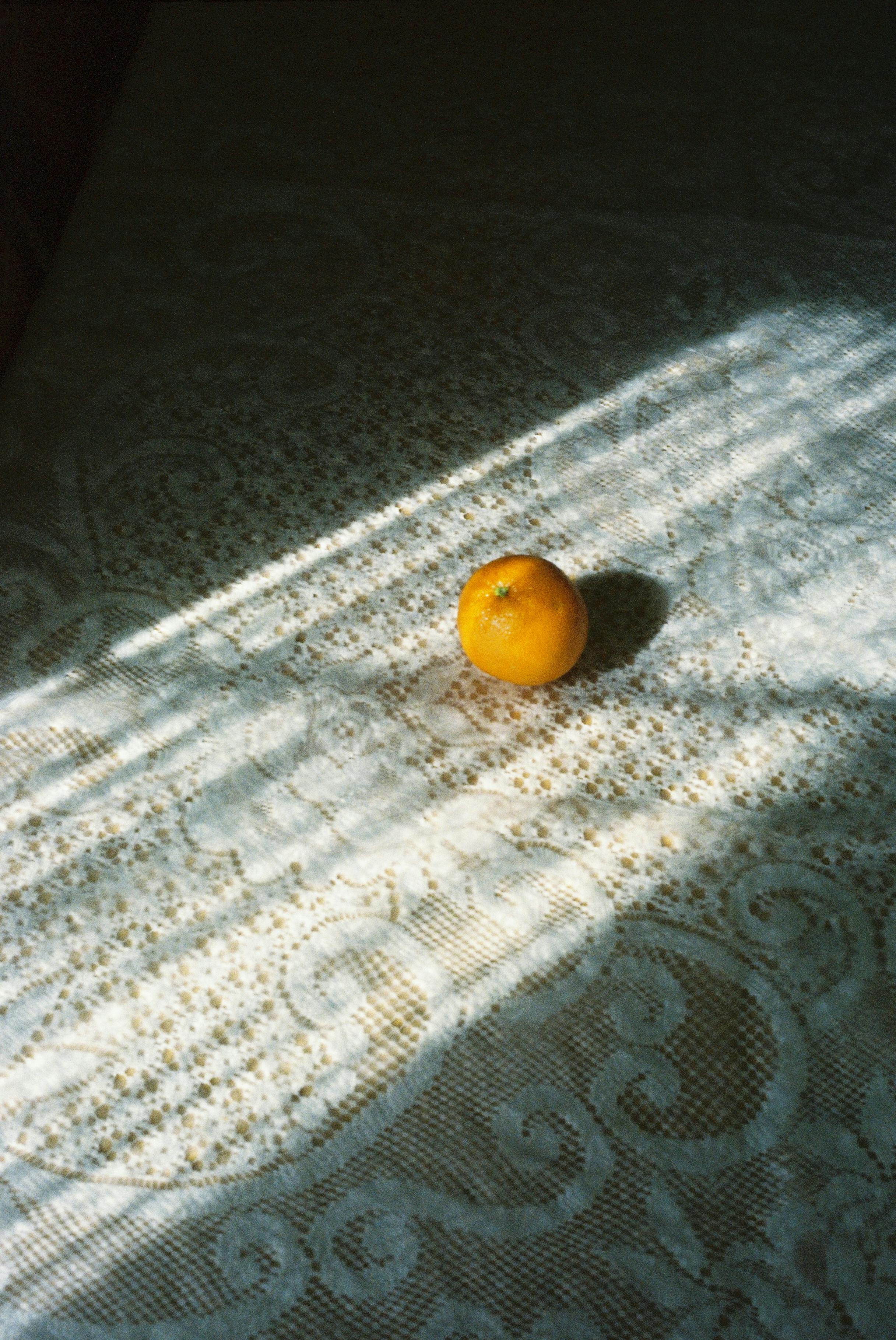 A lone tangerine sits in sunlight on a decorated lace tablecloth, evoking simplicity and warmth.