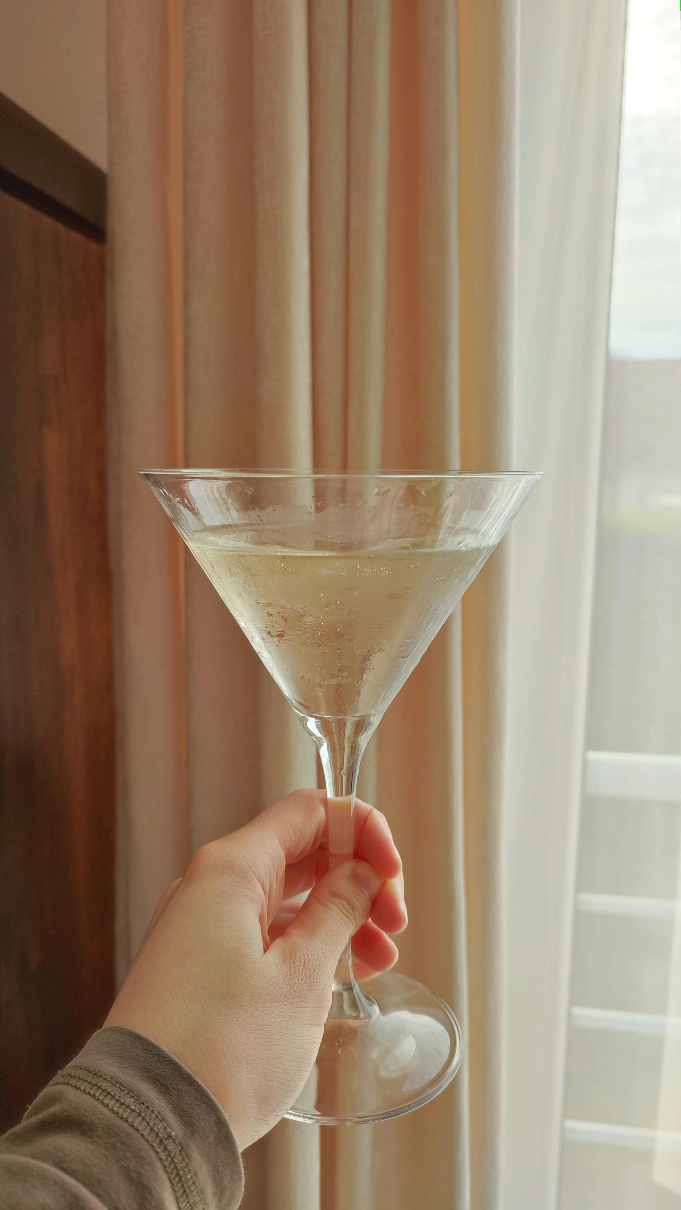 A hand holding a glass of champagne indoors against a bright curtain backdrop.