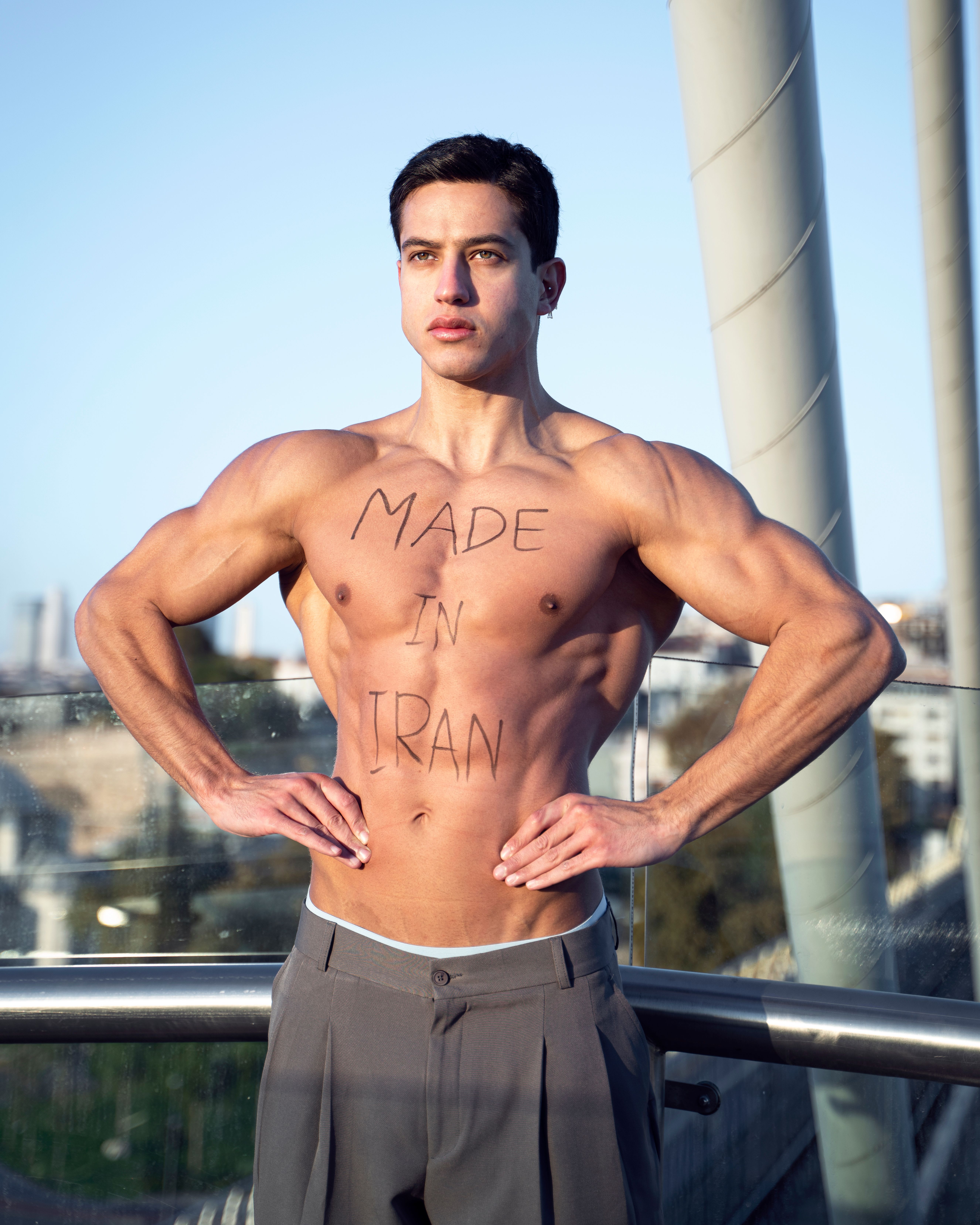 Shirtless muscular man flexing outdoors in Istanbul with 'Made in Iran' text on chest.