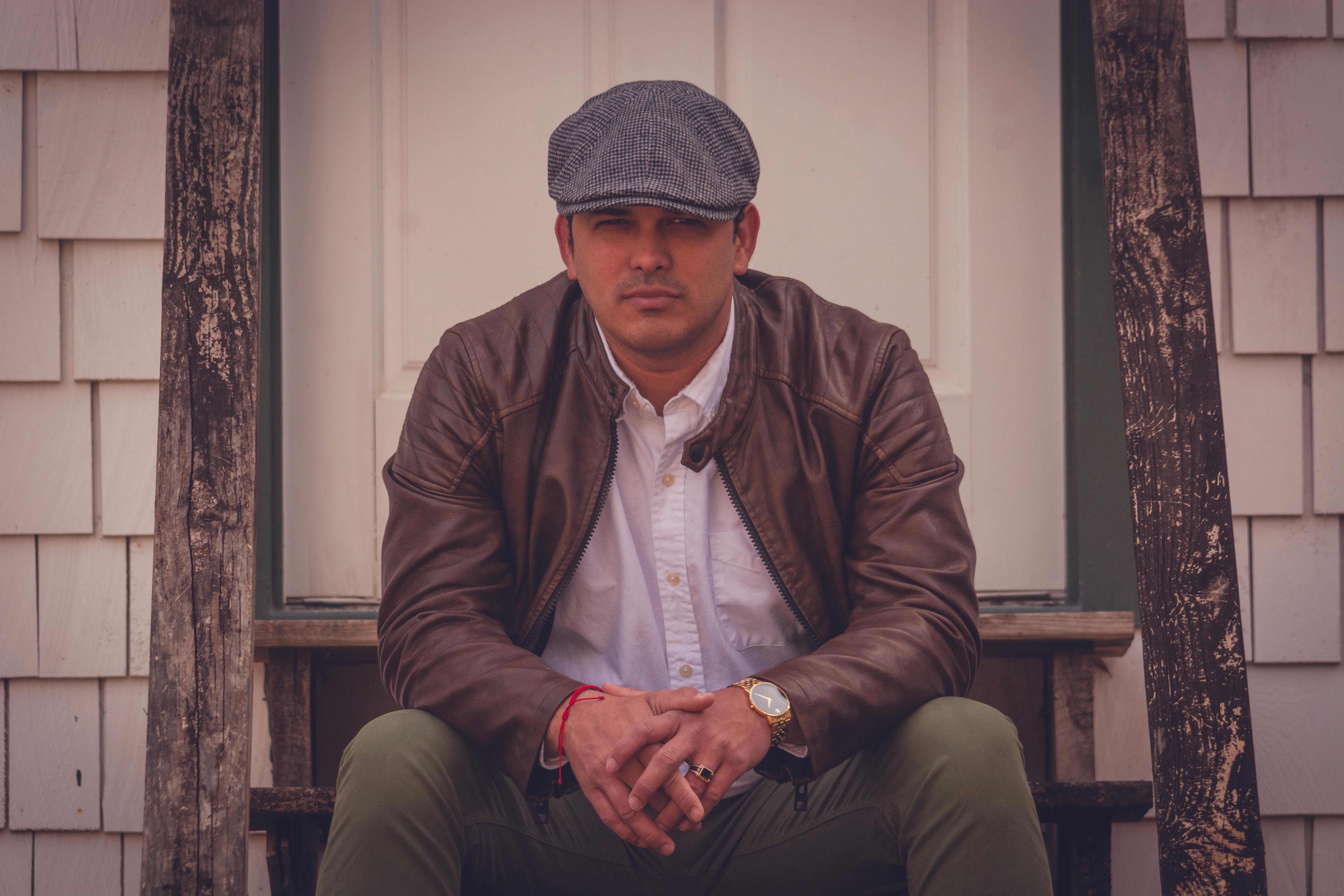 Man in leather jacket and cap sitting on vintage steps in a stylish pose.
