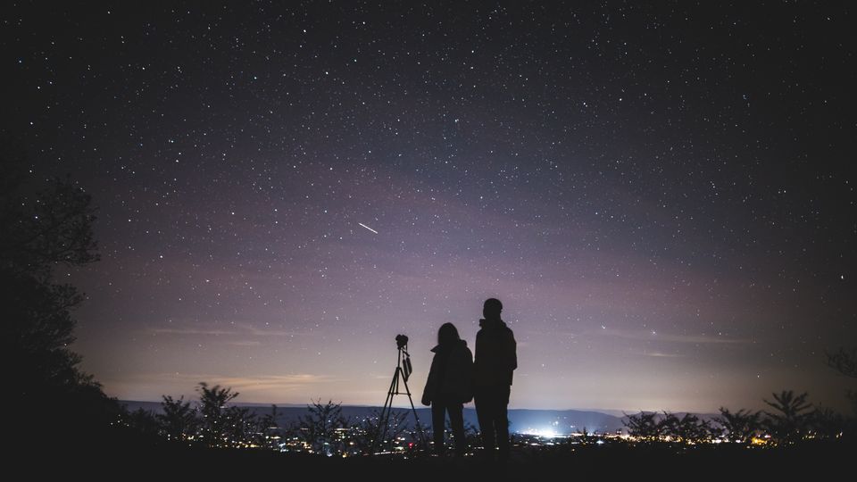 Two people stand beside a camera on a tripod on a hill under a starry evening sky