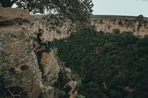 Free A man sitting on a rock cliff above a lush green canyon, enjoying the view. Stock Photo