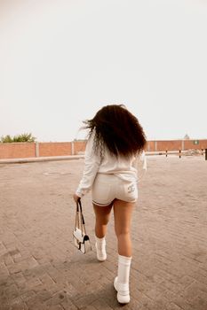 A woman with curly hair walks on a sunny urban sidewalk, showcasing a stylish summer outfit.