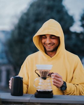 A cheerful man in a yellow hoodie brews coffee outdoors, enjoying a pleasant day in Eskişehir, Türkiye.