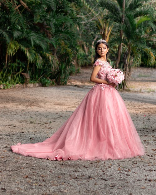 A Girl in a Pink Tulle Dress and a Tiara, Holding Flowers and Standing on the Background of Palm Trees 