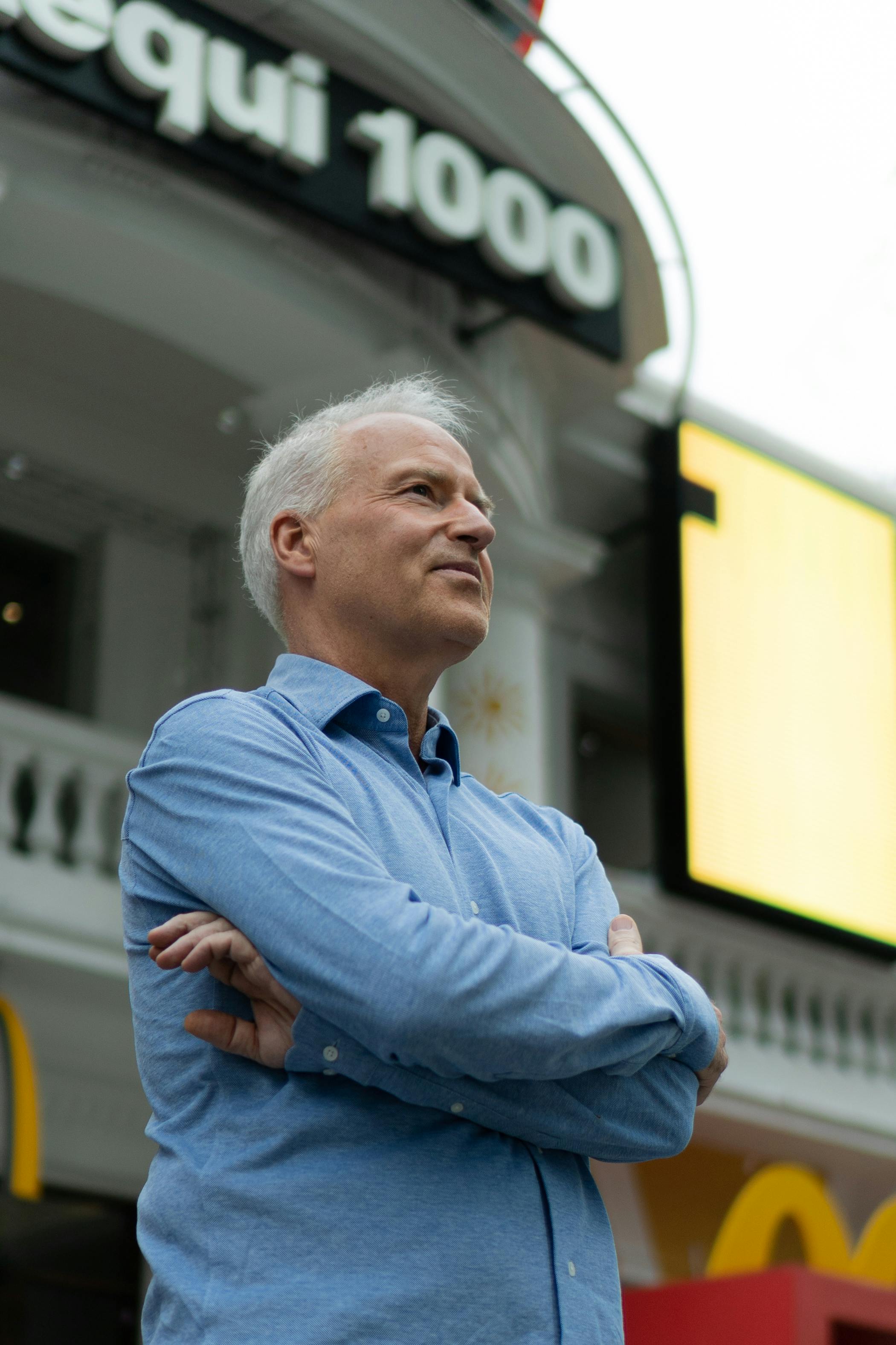 Senior man with gray hair and blue shirt stands confidently outdoors, arms crossed, below a building sign.