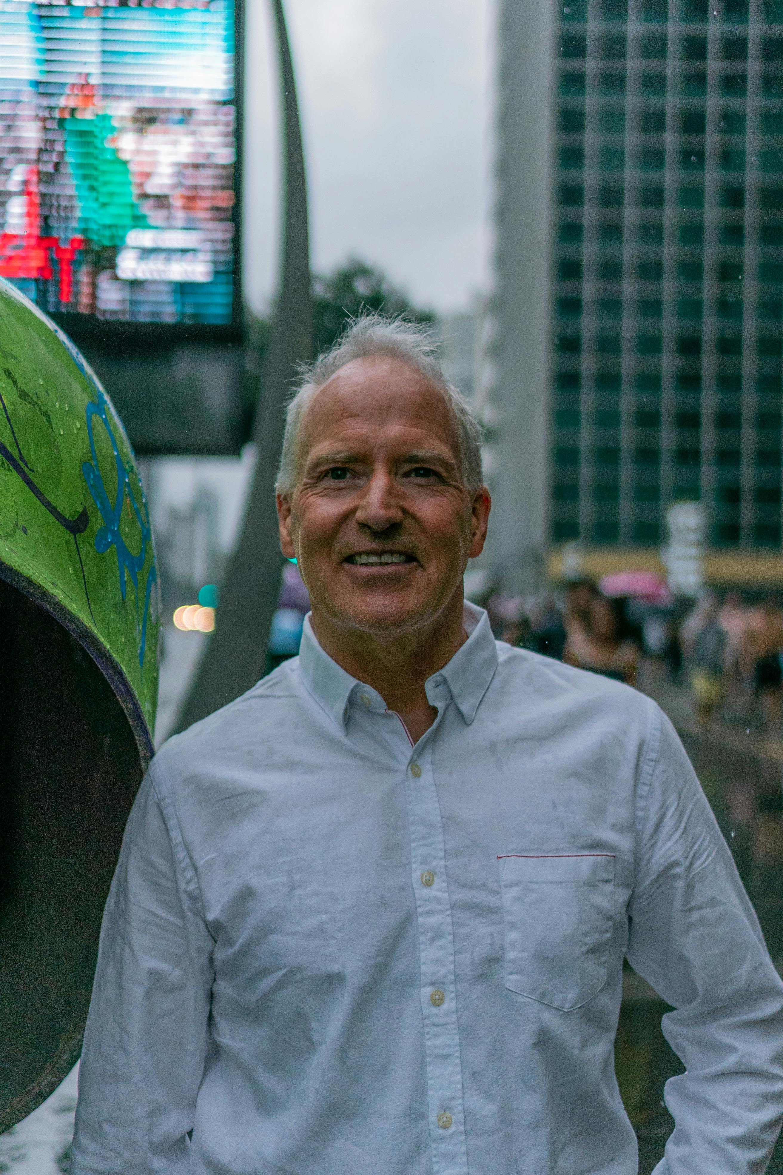 Portrait of a smiling man with gray hair in a city, wearing a white shirt, posing outdoors on a rainy day.