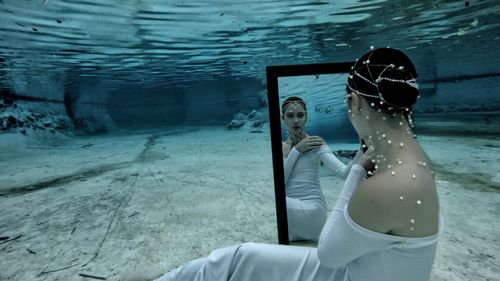 Free A woman in a white dress sits underwater holding a mirror, creating an ethereal reflection. Stock Photo