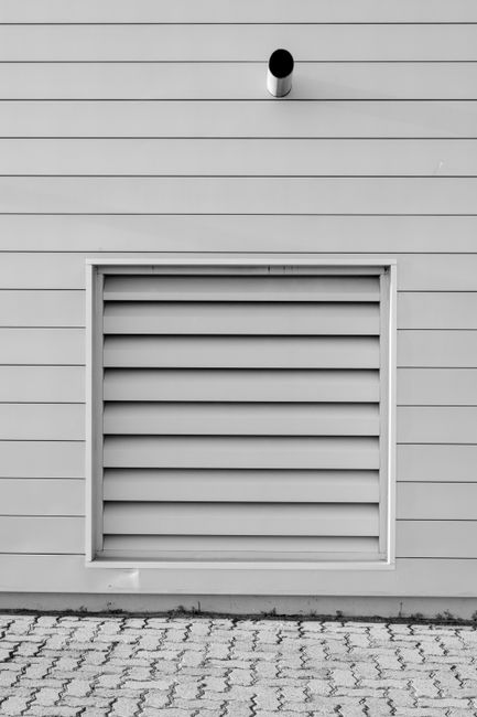 Black and white minimalist photo of an exterior wall with a vent and piping, capturing modern architecture.