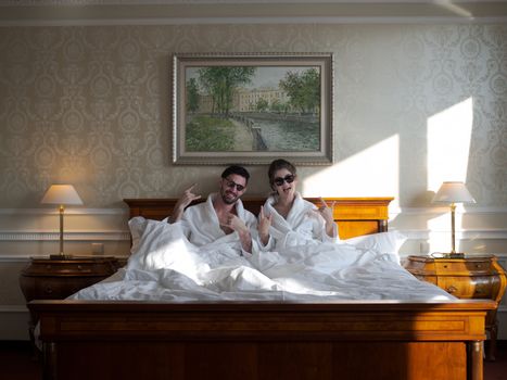 A couple wearing sunglasses and bathrobes relax in a stylish hotel room.