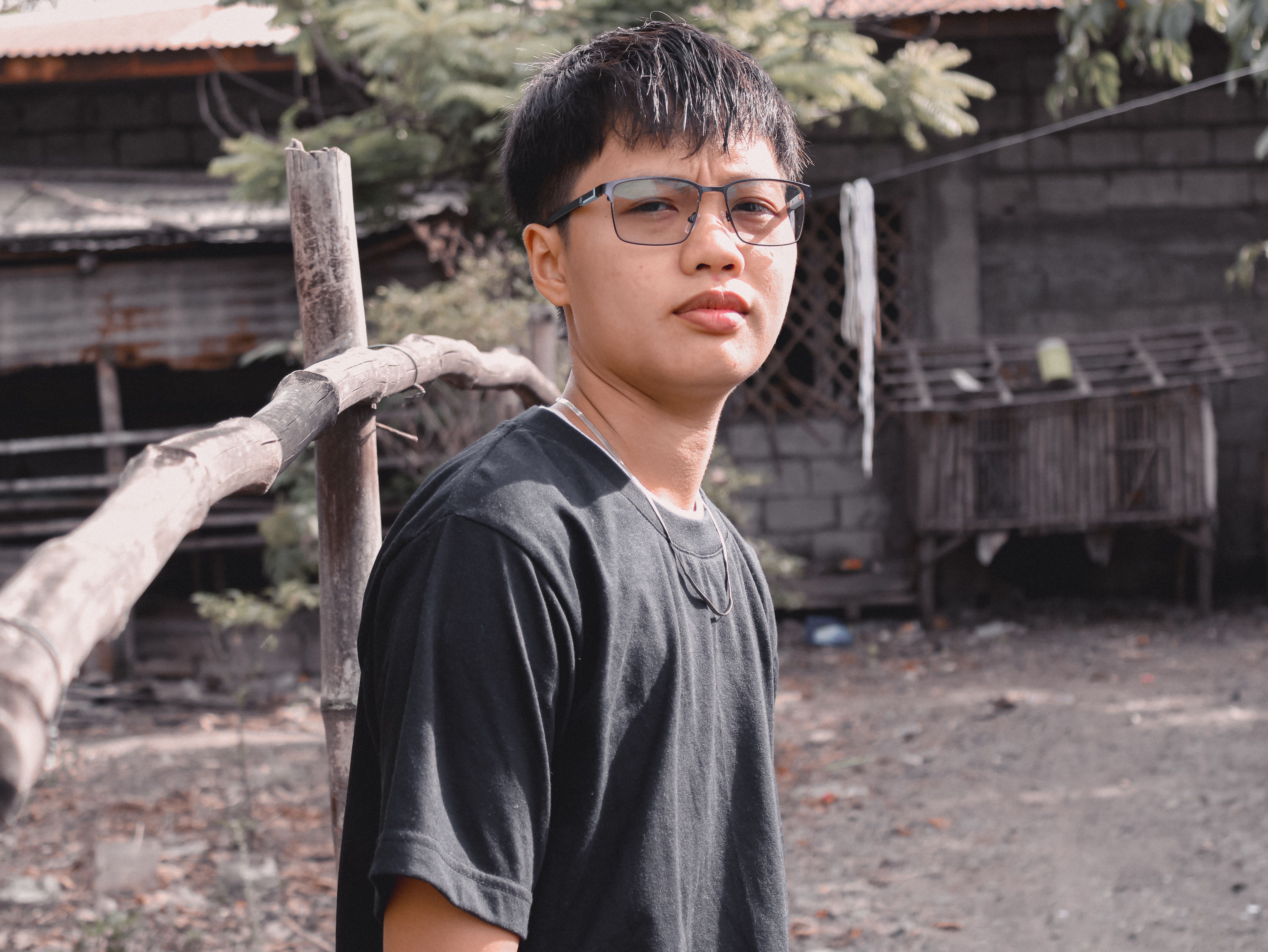 A young man wearing eyeglasses and a black t-shirt poses outdoors, embodying casual fashion.